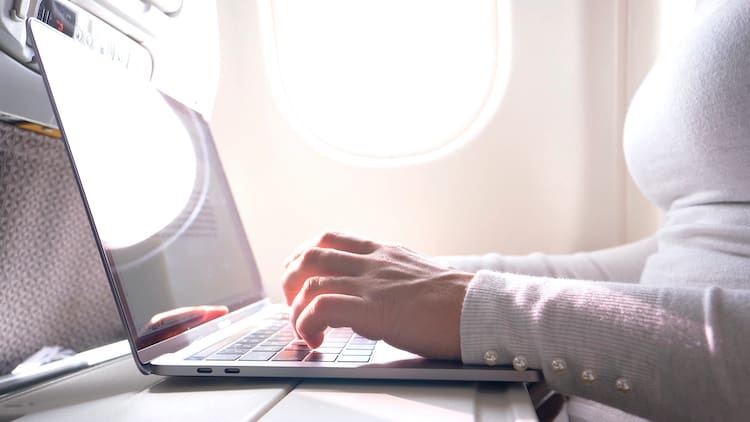 A woman on a plane with the window in the background. She is writing on her MacBook laptop.